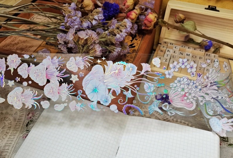 Puede incluir: Una hoja de cinta washi hologr&aacute;fica iridiscente con un dise&ntilde;o floral. La cinta presenta flores delicadas, hojas y remolinos en tonos de rosa, azul y blanco. La cinta es perfecta para agregar un toque de brillo y fantas&iacute;a a tus manualidades y proyectos.
