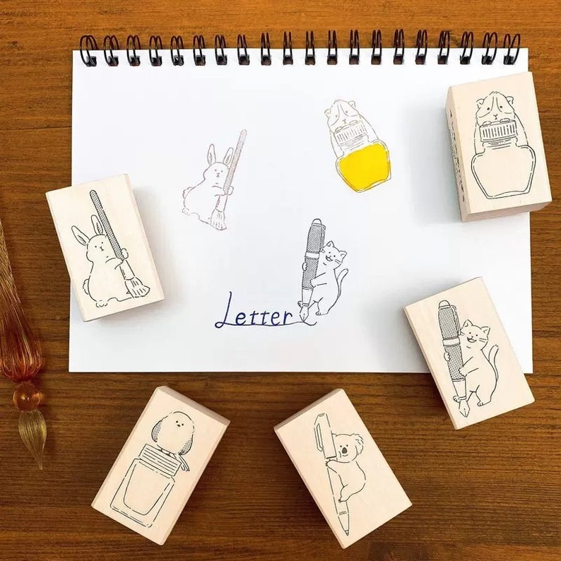 May include: Set of six wooden stamps featuring various animals holding pens and ink bottles. The stamps are on a white sheet of paper with the word "Letter" written in blue ink.