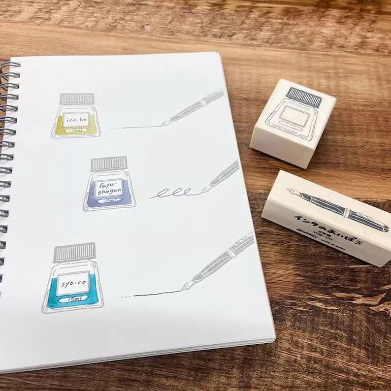 May include: Three ink bottles with labels reading "Ina-ho", "Fuyu Shogun", and "Syo-ro" are drawn on a white notebook page. A wooden stamp with the text "インクあそび" is shown next to the notebook. The stamp features a drawing of a fountain pen.