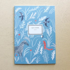 Puede incluir: Una libreta azul con un dise&ntilde;o caprichoso que presenta un caballo, una ardilla, p&aacute;jaros y elementos florales. La portada dice "NOTEBOOK".