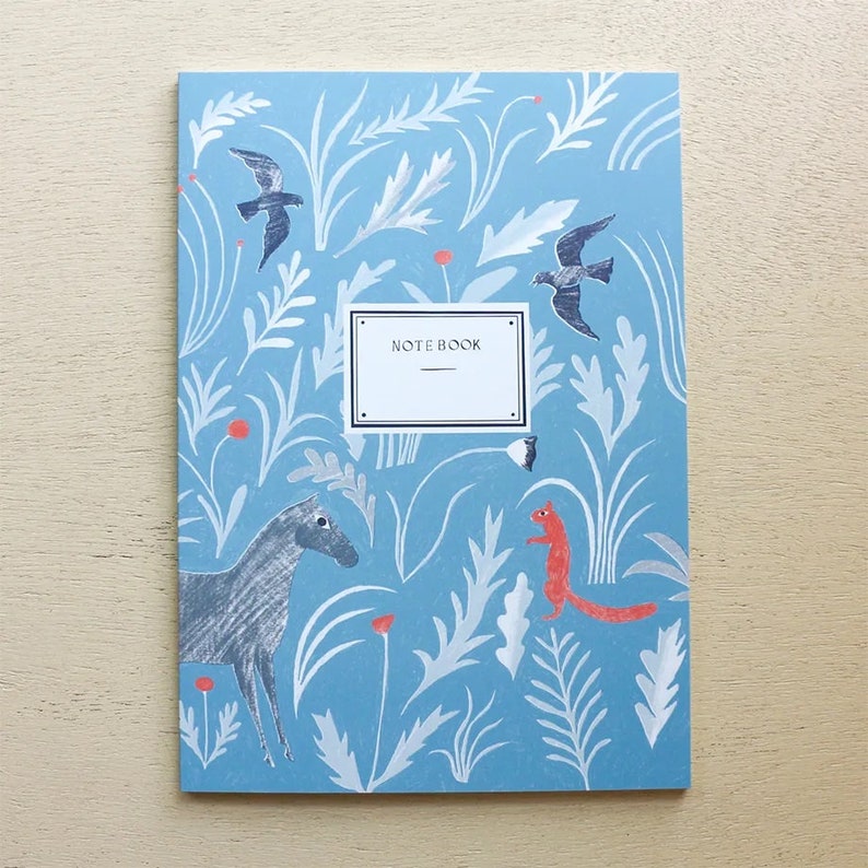 Puede incluir: Una libreta azul con un dise&ntilde;o caprichoso que presenta un caballo, una ardilla, p&aacute;jaros y elementos florales. La portada dice "NOTEBOOK".