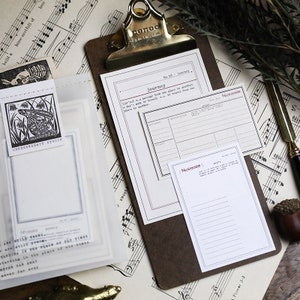 May include: A white printable planner page with the title "Journey" and a section for "Memory Book". The page is on a brown clipboard with a black and white musical note background.