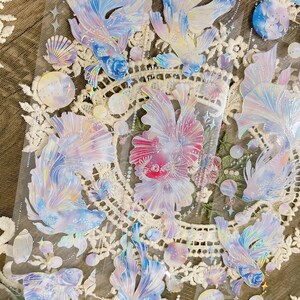 May include: A sheet of iridescent holographic stickers featuring various fish designs. The stickers are on a white background with a lace pattern. The fish are in shades of blue, pink, and purple.