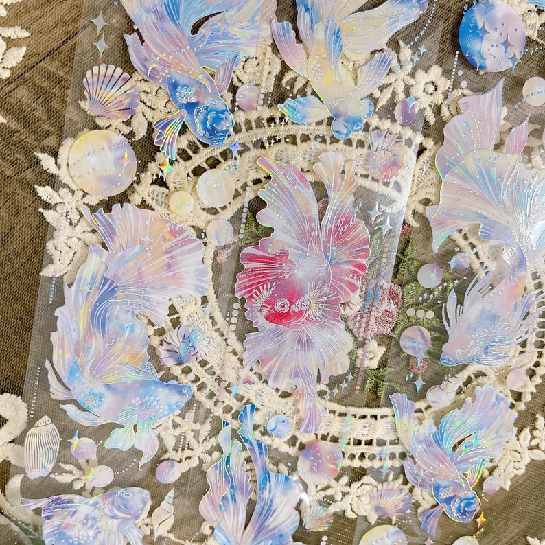 May include: A sheet of iridescent holographic stickers featuring various fish designs. The stickers are on a white background with a lace pattern. The fish are in shades of blue, pink, and purple.