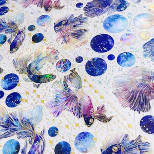 May include: A sheet of colorful stickers featuring iridescent blue, purple, and pink fish with galaxy-like patterns. The fish are surrounded by gold stars and blue and white galaxy-themed circles.