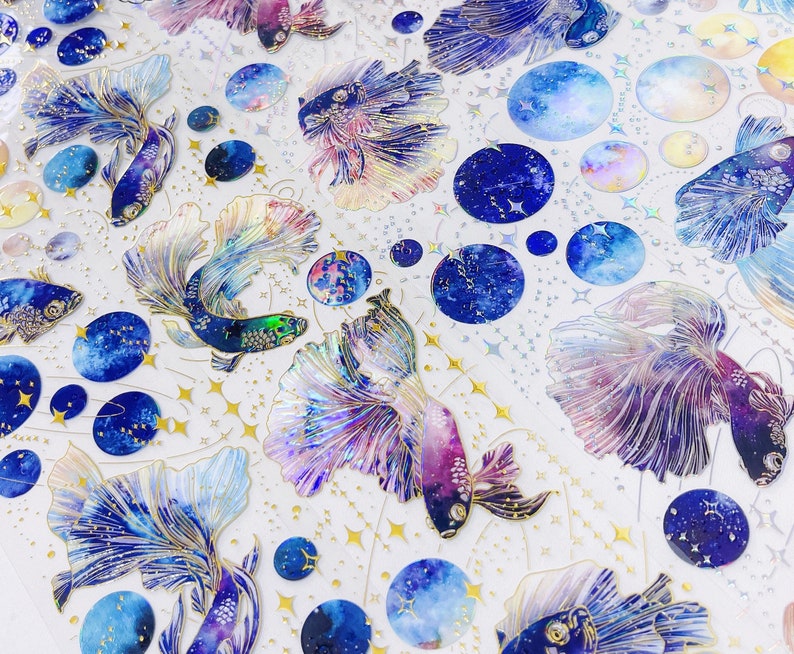 May include: A sheet of colorful stickers featuring iridescent blue, purple, and pink fish with galaxy-like patterns. The fish are surrounded by gold stars and blue and white galaxy-themed circles.
