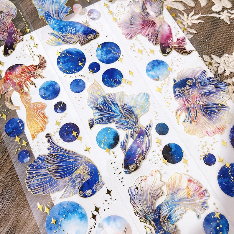 May include: A sheet of decorative stickers featuring colorful watercolor fish with galaxy-themed details. The fish have flowing fins and tails, and the galaxy designs include stars and swirls. The stickers are perfect for adding a touch of whimsy and magic to journals, planners, and other craft projects.