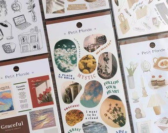 Op de afbeelding: Een set kleurrijke stickers met verschillende ontwerpen, waaronder bloemen, wolken, zonsondergangen en tekst. De stickers zijn gelabeld "Petit Monde" en bevatten zinnen als "Een plek om te ontspannen", "Mystiek", "Volg je hart" en "Ik wil een wolk zijn".