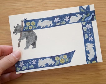 May include: A white card with a blue and white ribbon border. The ribbon is decorated with white rabbits, flowers, and green leaves. A black bear with a stick on its back is in the top left corner of the card.