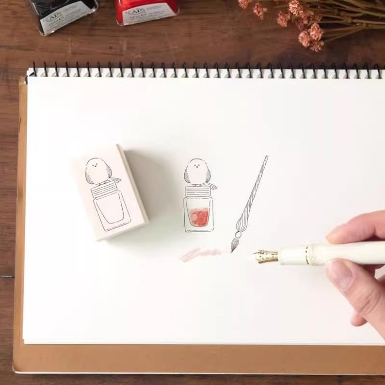 May include: Two wooden stamps with a bird perched on top of a bottle of ink. The stamps are on a white sheet of paper. A calligraphy pen is on the paper with a small amount of pink ink.
