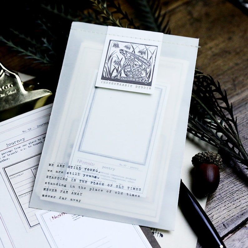 May include: A white translucent envelope with a black and white illustration of a rabbit in a field of flowers. The envelope has a black and white text overlay that reads "WE ARE STILL YOUNG, we are still young, poetry STANDING IN THE PLACE OF OLD TIMES NEVER FAR AWAY standing in the place of old times never far away" and "CHERSERABBIT STUDIO".