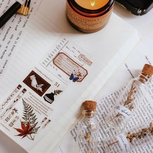 May include: A journal page with decorative elements. Includes botanical-themed washi tape with illustrations of leaves, test tubes, and a hand holding flowers. Small glass bottles with corks and dried plants are also present.