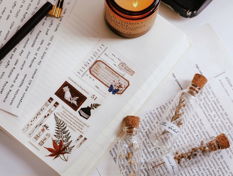 May include: A journal page with decorative elements. Includes botanical-themed washi tape with illustrations of leaves, test tubes, and a hand holding flowers. Small glass bottles with corks and dried plants are also present.