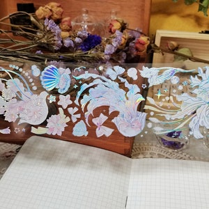 Puede incluir: Una hoja de pegatinas hologr&aacute;ficas iridiscentes con una variedad de dise&ntilde;os, incluyendo conchas marinas, flores y p&aacute;jaros. Las pegatinas son perfectas para agregar un toque de brillo a los diarios, planificadores y otros proyectos de manualidades.