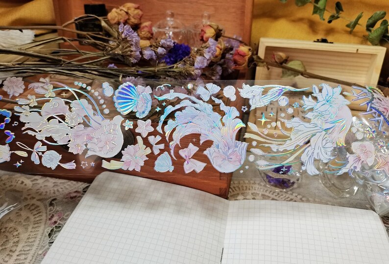 Puede incluir: Una hoja de pegatinas hologr&aacute;ficas iridiscentes con una variedad de dise&ntilde;os, incluyendo conchas marinas, flores y p&aacute;jaros. Las pegatinas son perfectas para agregar un toque de brillo a los diarios, planificadores y otros proyectos de manualidades.