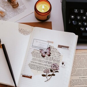 May include: An open notebook with lined pages, decorated with butterfly and floral stickers, and torn paper. A lit candle in a glass jar, a black pen, and a vintage typewriter are also visible.