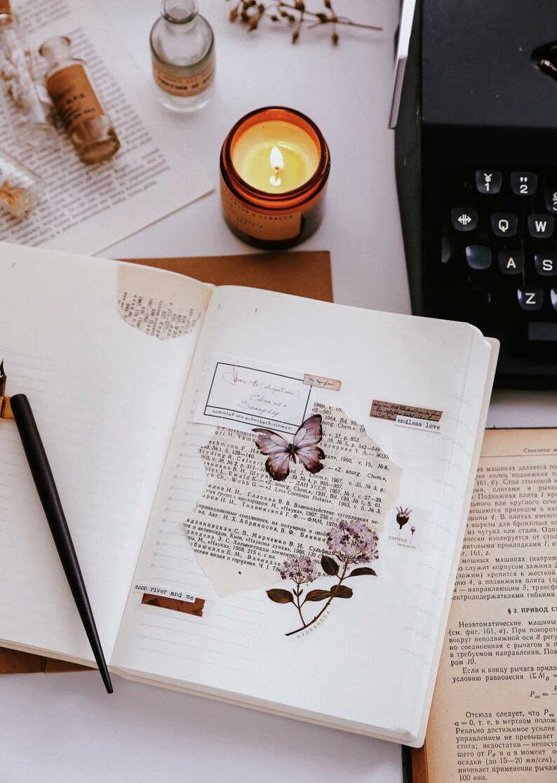 May include: An open notebook with lined pages, decorated with butterfly and floral stickers, and torn paper. A lit candle in a glass jar, a black pen, and a vintage typewriter are also visible.
