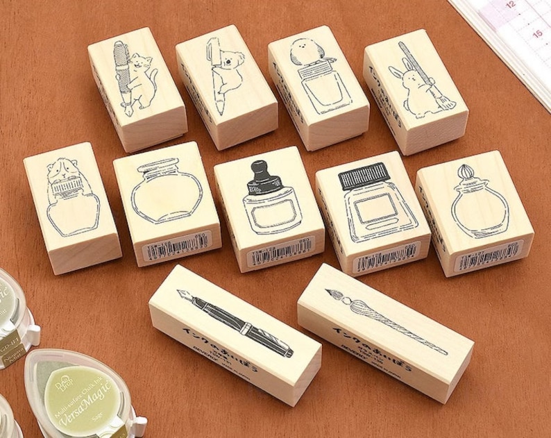 May include: A set of ten wooden stamps featuring various illustrations, including a cat, a koala, a bird, a rabbit, a bottle of ink, a pen, and a quill. The stamps are all rectangular and have a light brown color.
