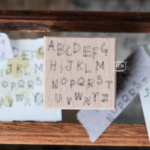 May include: A wooden alphabet stamp with uppercase letters A through Z in a handwritten style. The stamp is square and sits on a wooden surface, with examples of stamped letters on paper in the background.