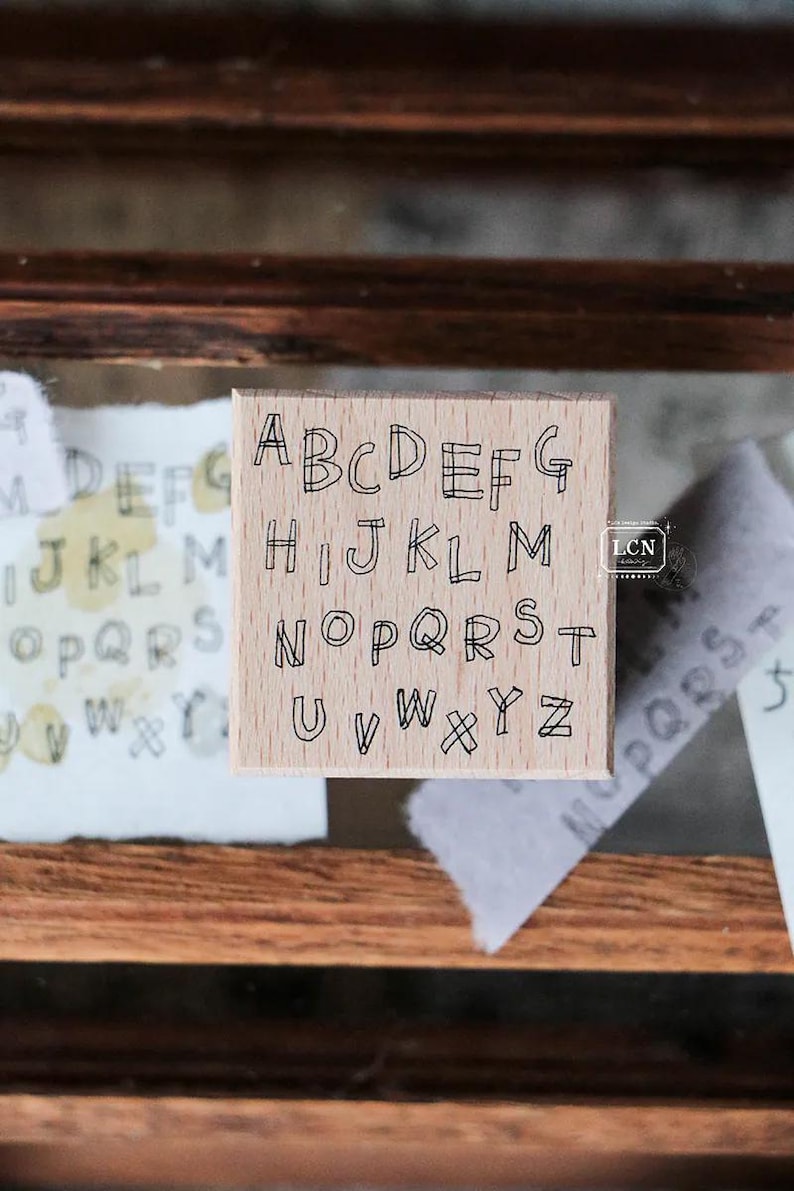 May include: A wooden alphabet stamp with uppercase letters A through Z in a handwritten style. The stamp is square and sits on a wooden surface, with examples of stamped letters on paper in the background.