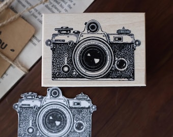 May include: A black and white camera design on a wooden stamp, with a smaller sticker version below. The stamp is rectangular and sits on a wooden surface. The design is a detailed illustration.