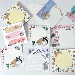 May include: A collection of square sticky notes with various designs featuring rabbits, flowers, and strawberries. Some notes have blank spaces for writing, while others have illustrations and the word "Rabbit". The color palette includes pink, white, and yellow.