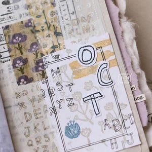 May include: A collage of vintage ephemera with floral and alphabet designs. The composition includes layered paper, stamps, and decorative tape. The color palette features muted tones of beige, yellow, and purple.