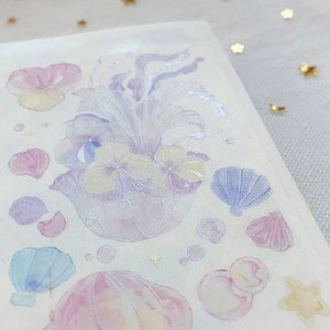 Puede incluir: Una hoja de pegatinas de color pastel con una medusa, flores, conchas marinas y otros dise&ntilde;os de tem&aacute;tica oce&aacute;nica. Las pegatinas son transl&uacute;cidas y tienen un acabado brillante.