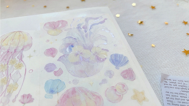 Puede incluir: Una hoja de pegatinas de color pastel con una medusa, flores, conchas marinas y otros dise&ntilde;os de tem&aacute;tica oce&aacute;nica. Las pegatinas son transl&uacute;cidas y tienen un acabado brillante.