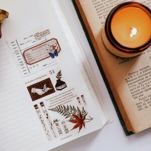 May include: An open notebook with decorative stickers, a wooden stamp, and a lit candle. The stickers feature botanical illustrations, a registration label, and a butterfly. The candle is in a glass jar.