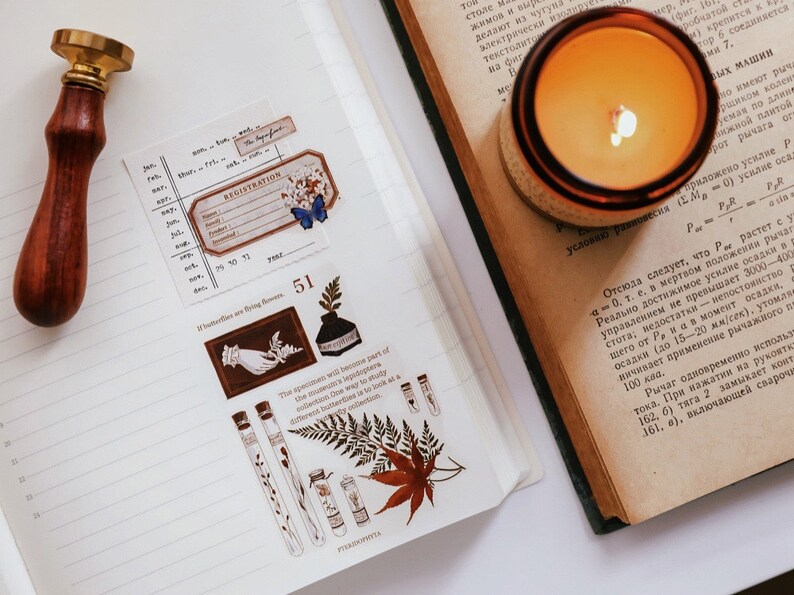 May include: An open notebook with decorative stickers, a wooden stamp, and a lit candle. The stickers feature botanical illustrations, a registration label, and a butterfly. The candle is in a glass jar.