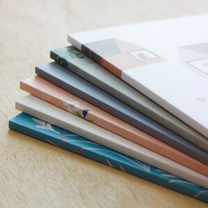 Puede incluir: Una pila de seis cuadernos con diferentes cubiertas estampadas. El cuaderno de arriba tiene una cubierta blanca con una ilustraci&oacute;n de una casa y el texto "Our Home". Los otros cuadernos tienen cubiertas con estampados florales, de hojas y geom&eacute;tricos en tonos azules, verdes, rosas y blancos.