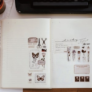 May include: Open notebook with decorative stickers featuring butterflies, flowers, and botanical elements. Small glass bottles with dried flowers and a lit candle are also visible, creating a vintage aesthetic.