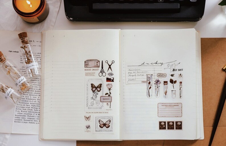 May include: Open notebook with decorative stickers featuring butterflies, flowers, and botanical elements. Small glass bottles with dried flowers and a lit candle are also visible, creating a vintage aesthetic.