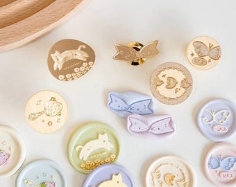Può includere: Assortimento di sigilli di cera colorati con vari disegni, tra cui gatti, farfalle, fragole e fiocchi. I sigilli sono fatti di cera e hanno una finitura dorata metallizzata.