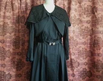 Op de afbeelding: Een zwarte vintage jurk met een kanten kraag en een decoratieve riem. De jurk heeft lange mouwen en een zwierige rok. De riem heeft een rechthoekige gesp met strass-steentjes. De jurk wordt gepresenteerd op een mannequin.