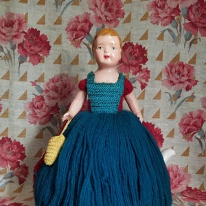 May include: A crocheted teapot cozy in the shape of a doll wearing a teal dress, red bodice, and a yellow crocheted bag. The doll has a porcelain face with painted features.