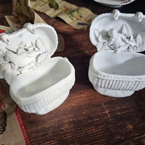May include: Two white ceramic basket-shaped trinket boxes with hinged lids. Each box features a woven basket design and a detailed relief of a bird and foliage inside the lid. The boxes are open, revealing the interior.