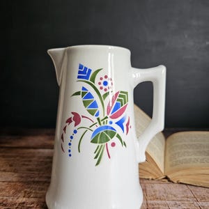 May include: White ceramic pitcher with a stylized floral design in shades of blue, green, red, and white. The pitcher has a wide base and a narrow spout.