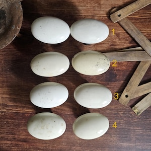 May include: A collection of six antique white oval cabinet knobs. The knobs are arranged in two rows of three, with varying shades of white and off-white. The knobs are on a dark wood surface, with a metal object and a wooden ruler in the background.