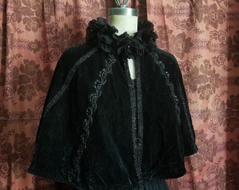 May include: A black velvet cape with beaded trim and a ruffled collar. The cape is displayed on a mannequin against a floral patterned background.