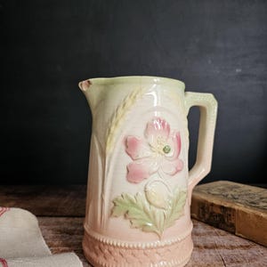 May include: A vintage ceramic pitcher with a floral design. The pitcher is decorated with a pink flower, green leaves, and wheat stalks. The pitcher has a light green glaze and a pink base.