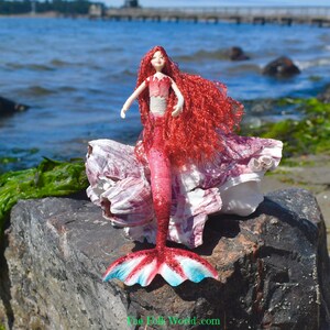 Puede incluir: Una muñeca de sirena hecha a mano con cabello rojo y una cola a juego, posada sobre una roca cerca del océano. La cola de la sirena presenta detalles en rojo, blanco y azul. La muñeca lleva un top rosa y está colocada sobre una gran concha.