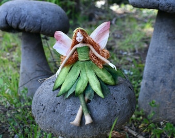 May include: A handcrafted fairy doll with red hair, a green dress, and leaf-like skirt sits atop a mushroom-shaped stone. The fairy has pink and white wings. The background features other mushroom-shaped stones and greenery.
