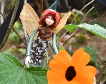 May include: A handcrafted fairy doll with a red cap and wings, set against a backdrop of greenery and an orange flower. The doll has a brown bodice and a skirt with a white floral pattern. The fairy is a whimsical, decorative item.