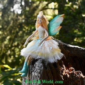 May include: A handcrafted fairy doll with blonde hair, a jeweled crown, and turquoise wings. The fairy is wearing a white and yellow dress with a sparkling bodice and turquoise boots. The doll is sitting on a weathered wooden surface.