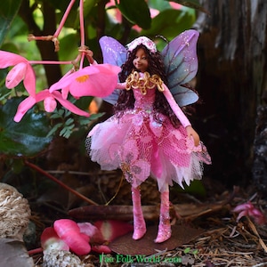 Puede incluir: Una muñeca de hada en miniatura con cabello castaño, vestida con un vestido rosa con una falda en capas y alas brillantes. El hada sostiene una flor rosa, sobre un fondo natural de vegetación y flores.