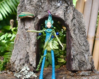 May include: A whimsical fairy figurine with a blue and green color scheme. The fairy has a pointed hat, outstretched arms, and is adorned with blue orbs. It stands in front of a miniature tree trunk with a doorway, surrounded by greenery.