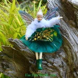 May include: A miniature fairy doll with a white hat and a white fluffy shawl. The fairy has green and gold accents on its dress and boots. The fairy has translucent wings with blue and purple hues. The text "Fae Folk World.com" is at the bottom.