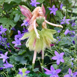 May include: A fairy doll with blonde hair, a green bodice, and a layered green skirt with pink accents. The doll has pink and purple wings and is surrounded by purple flowers and green foliage. The doll is approximately 15 cm tall.